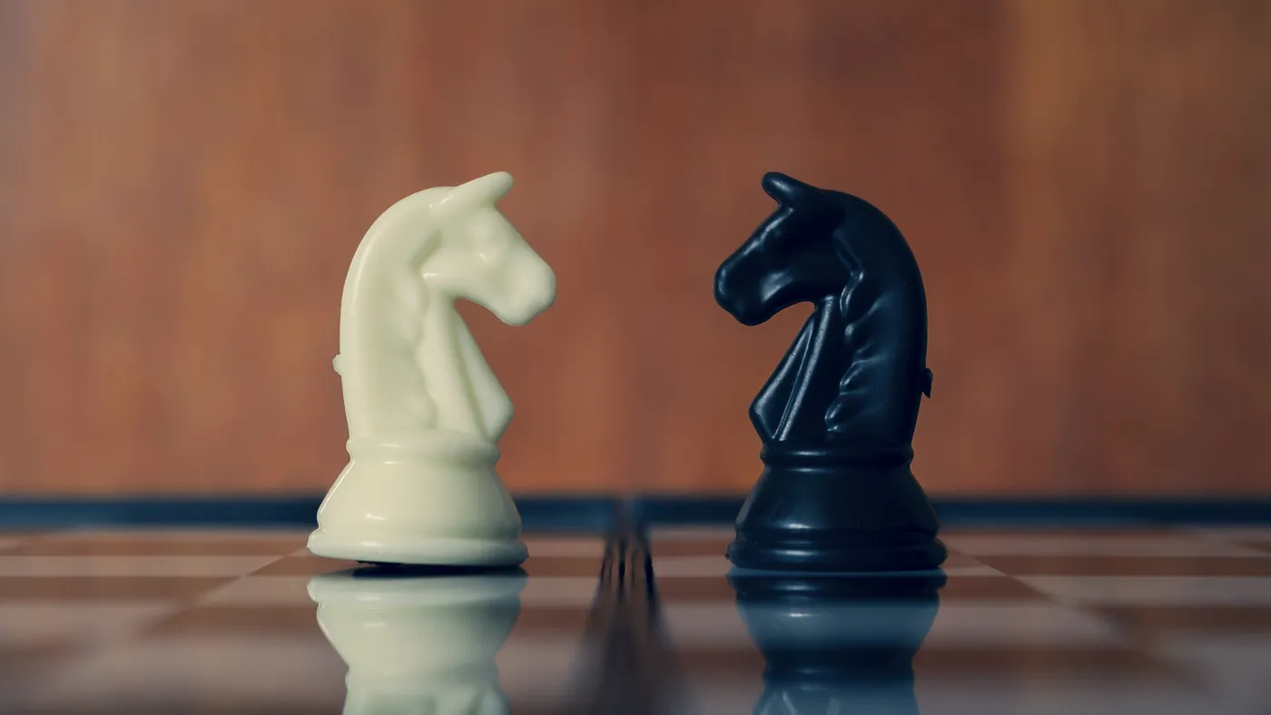 A white chess knight and a black chess knight face each other on a glossy chessboard