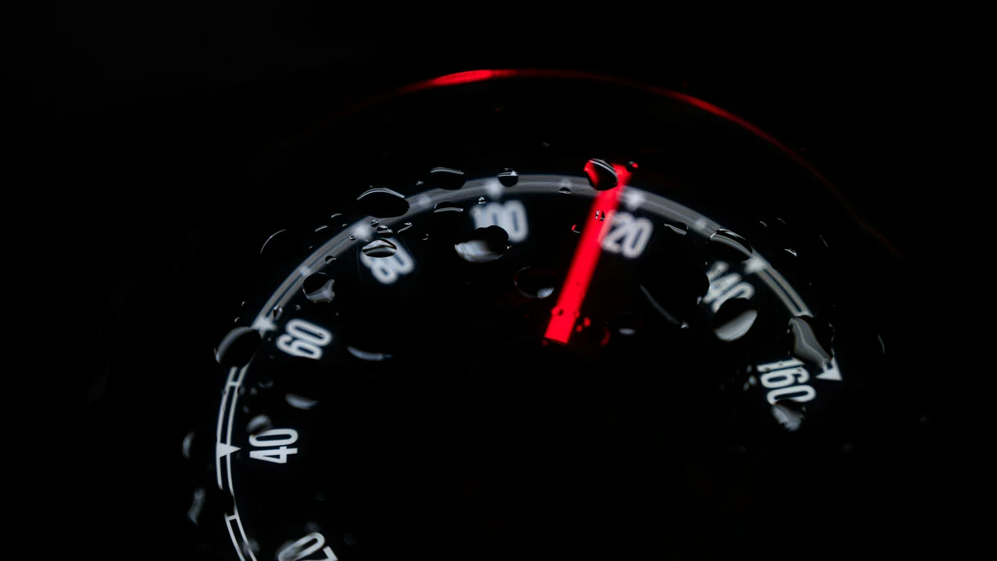 A rain-speckled speedometer in near darkness, its red needle swung past 100 and still climbing