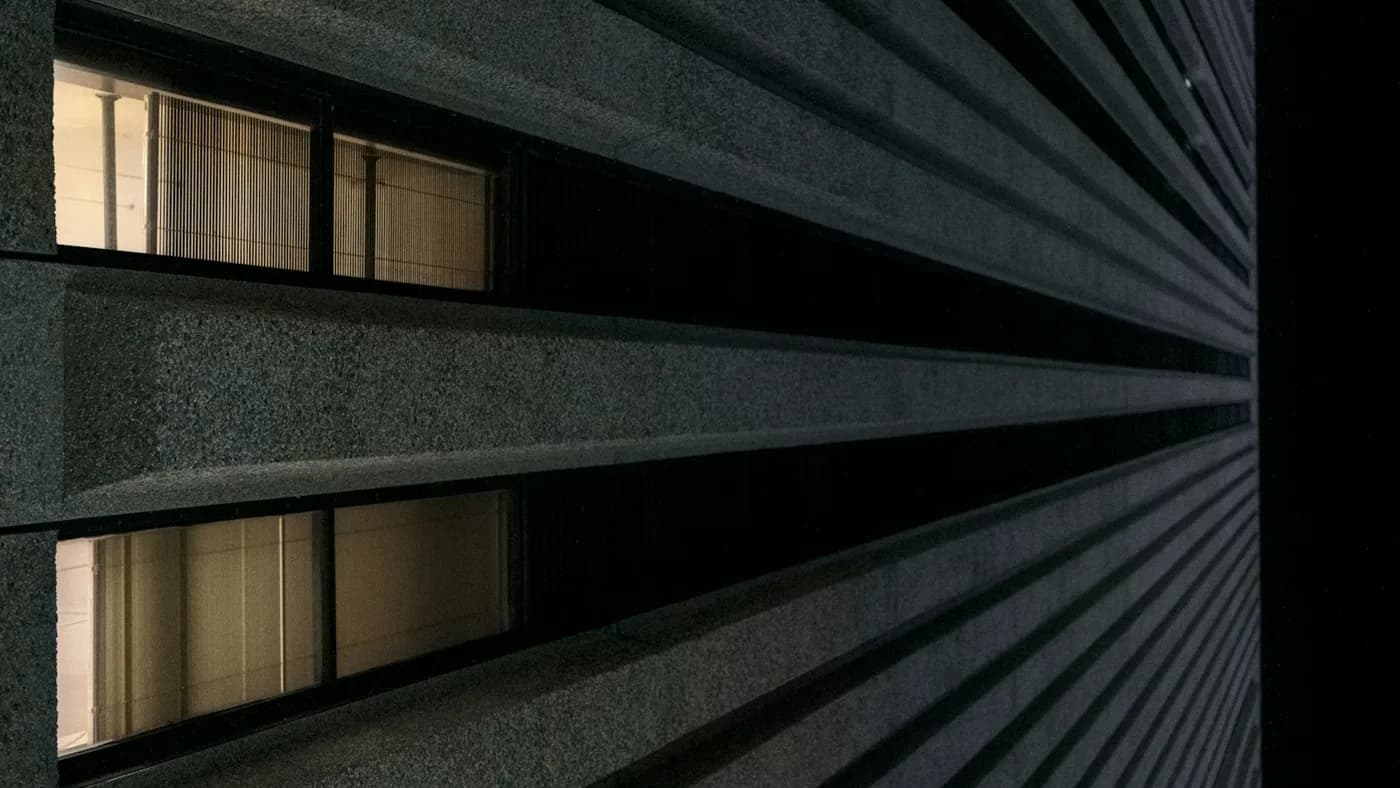 Dark concrete facade at night with parallel horizontal ledges receding past two lit windows