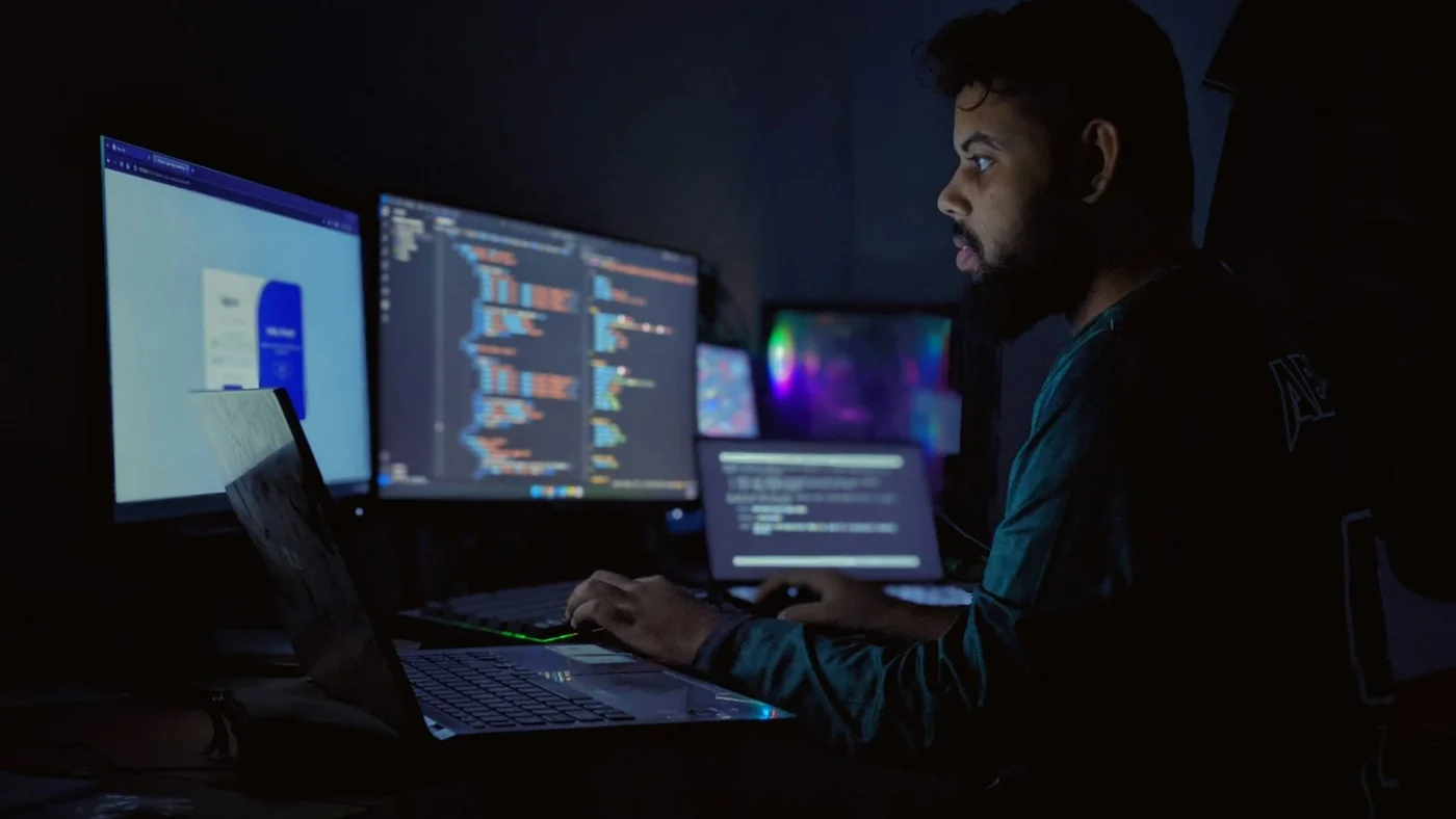 Developer working at multiple monitors in a dark room with code and AI output on screen