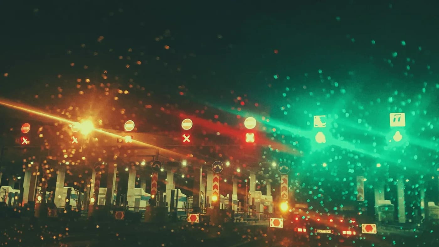 Rain-streaked windshield view of a lit toll plaza at night, red and green lane signals in a row