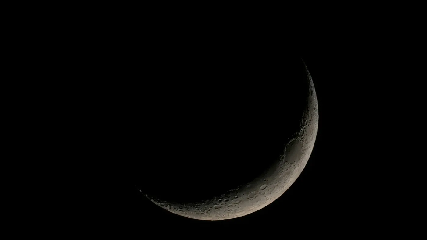 Thin crescent moon against a black night sky, with craters visible along its lit lower edge