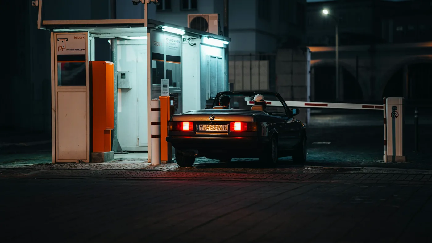A car waits at a lit parking payment booth at night with the striped barrier arm down across the lane