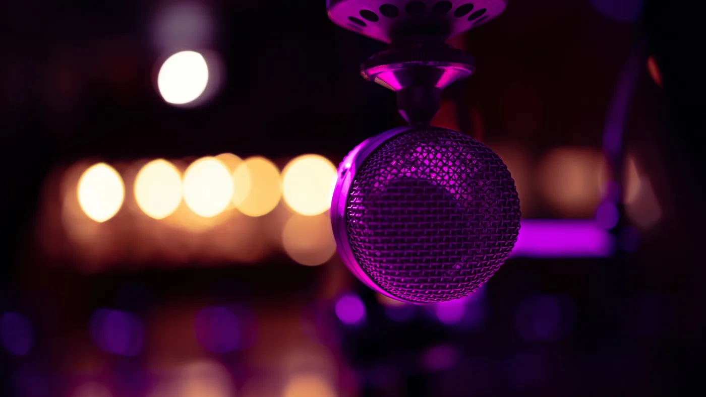Close-up of a hanging studio microphone lit purple in a dark room with warm bokeh lights behind it
