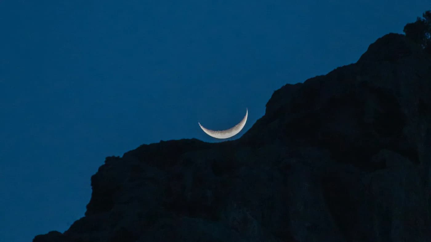 Thin crescent moon resting on a dark rocky ridge against a deep blue night sky