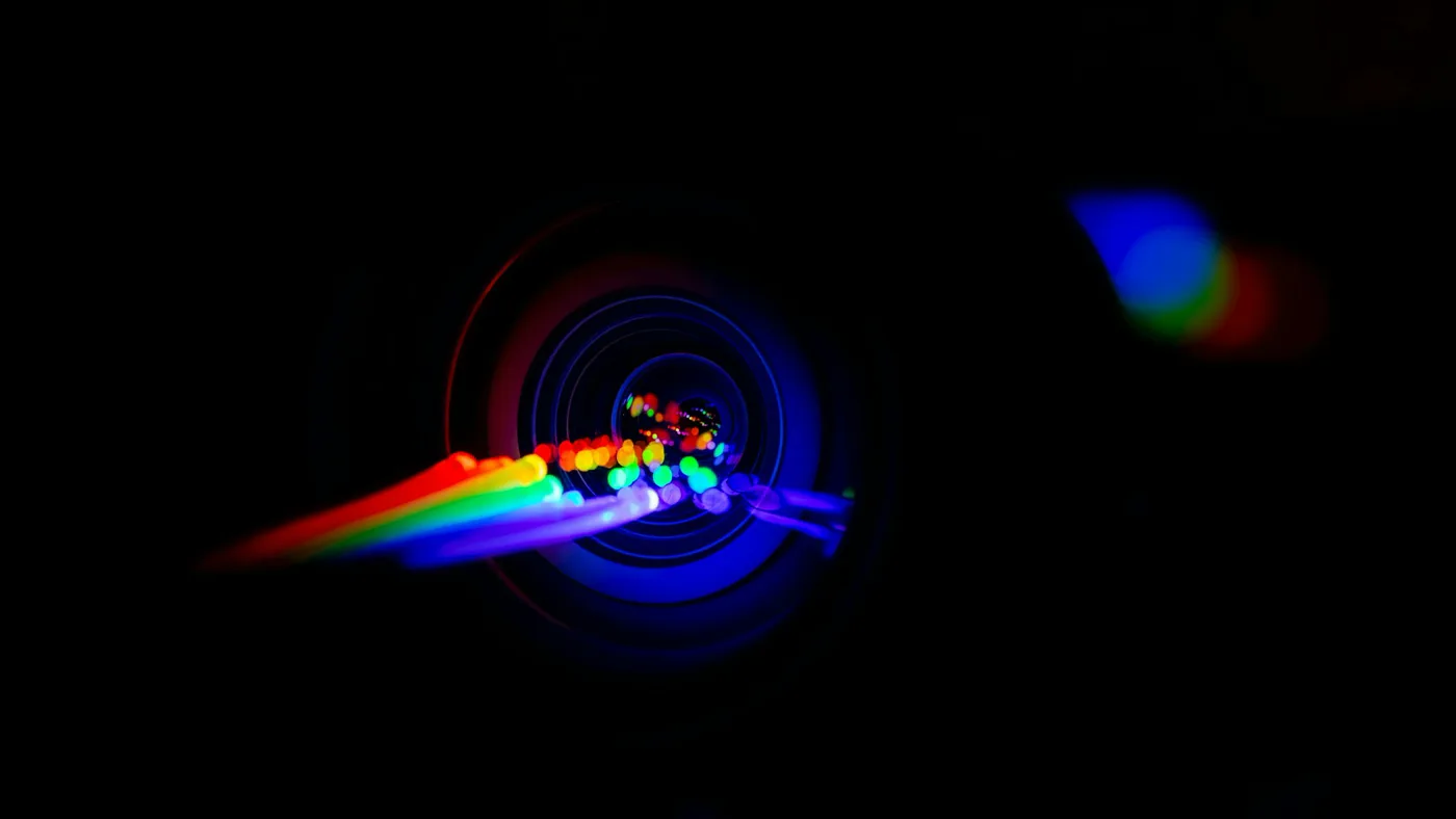 A rainbow streak of light running into a glowing blue camera lens on a black background