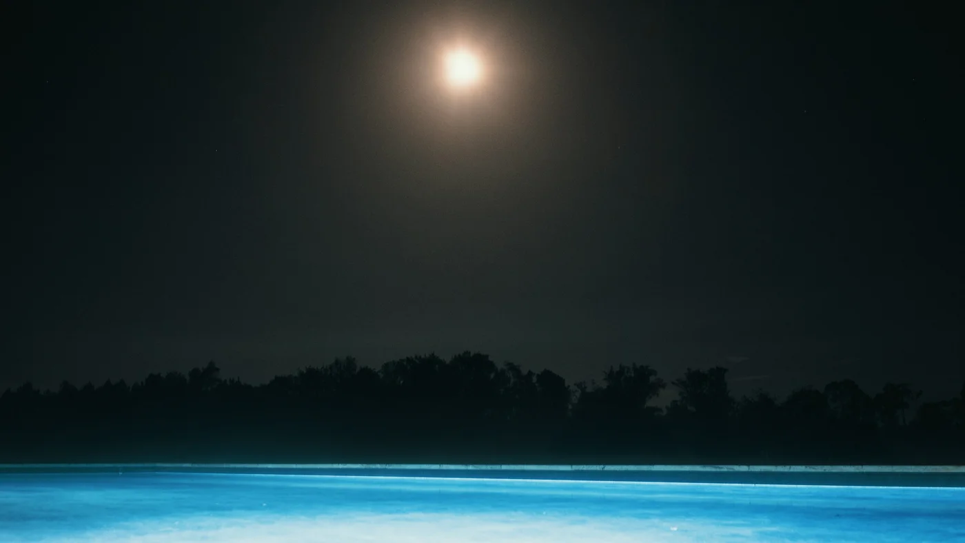 Moonlit night sky over a glowing blue swimming pool with a dark tree line