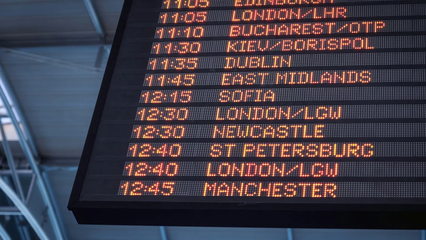 Airport departure board with amber LED times and destinations, seen from below against a dim steel roof