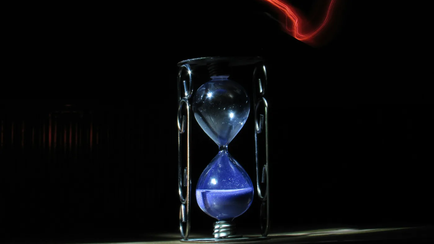 Glass hourglass with blue sand in a metal stand on a dark table, a red light trail curving through the black